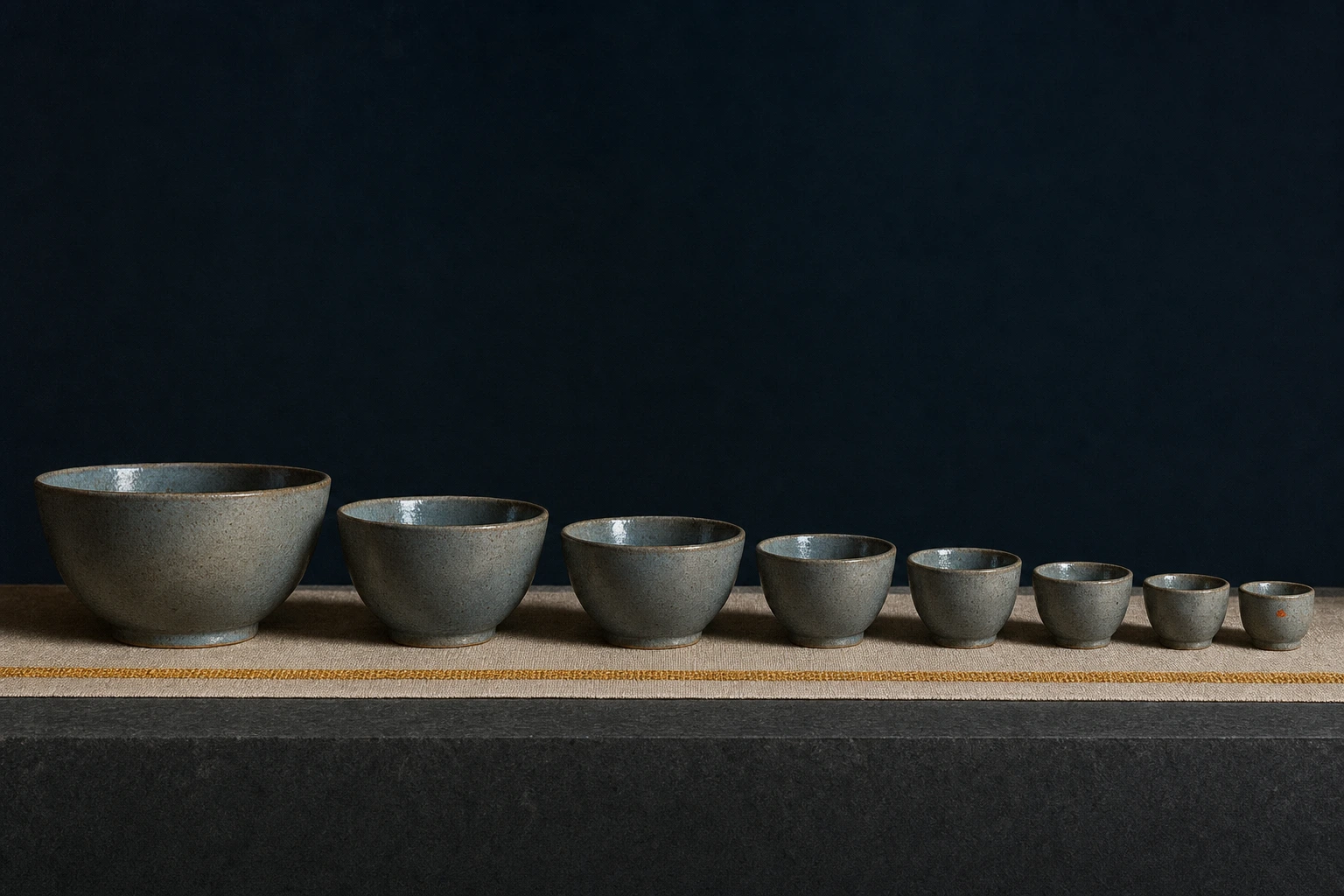 A procession of stoneware bowls steps down evenly in size along a dark bench on a narrow linen runner with a warm gold hem.