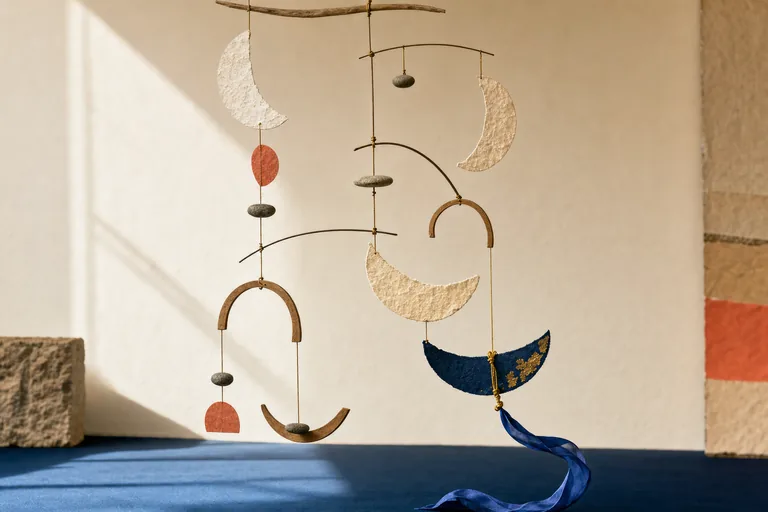 Paper arcs and river stones rise and fall in a balanced mobile as a knotted gold thread releases.